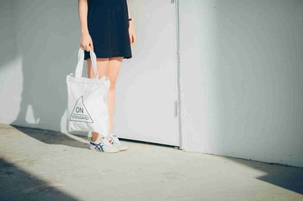 Seorang wanita mengenakan gaun hitam dan sepatu kasual, memegang totebag putih di depan tembok putih